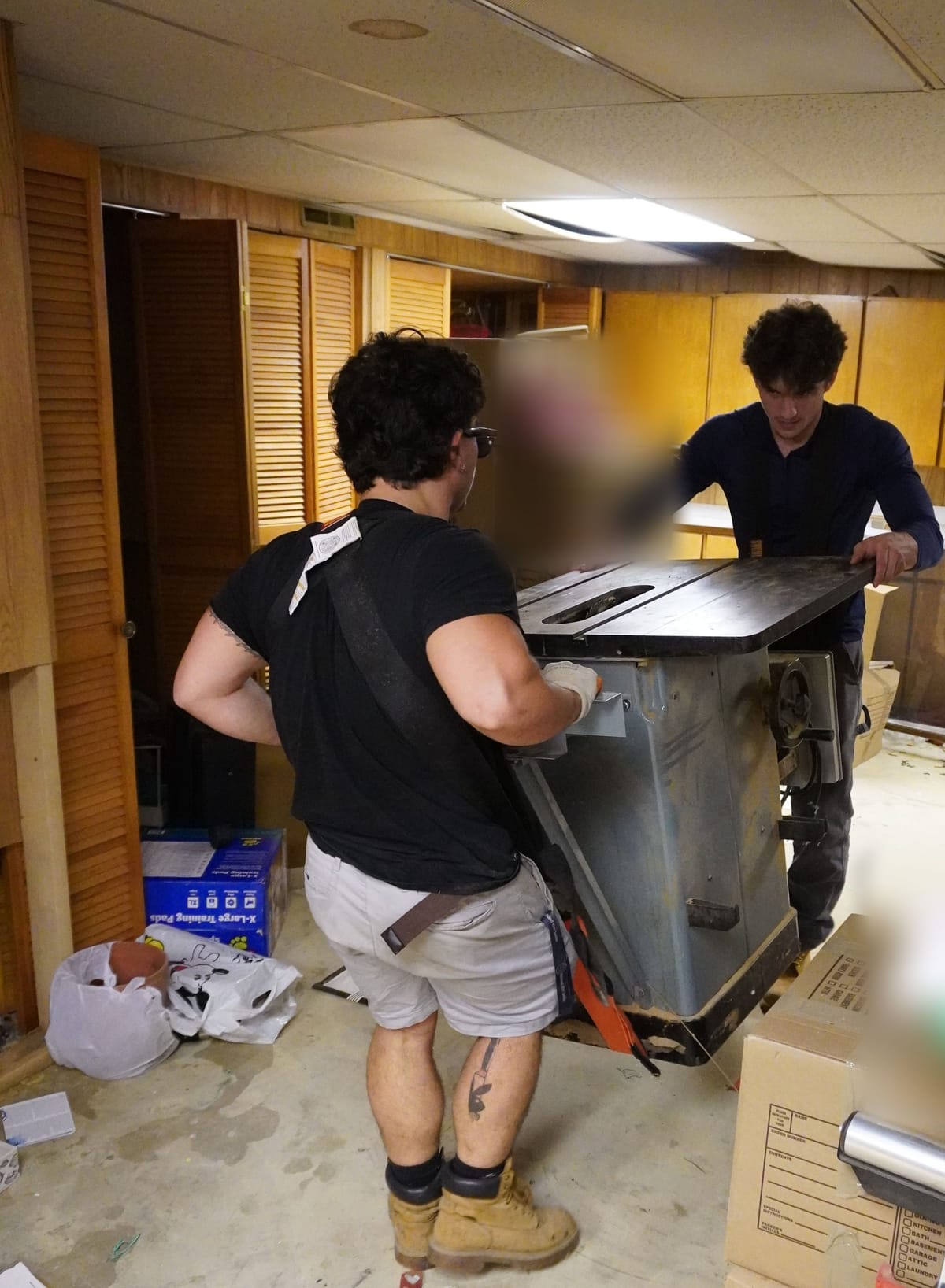 Two Safe Move movers carrying a heavy table saw across a basement